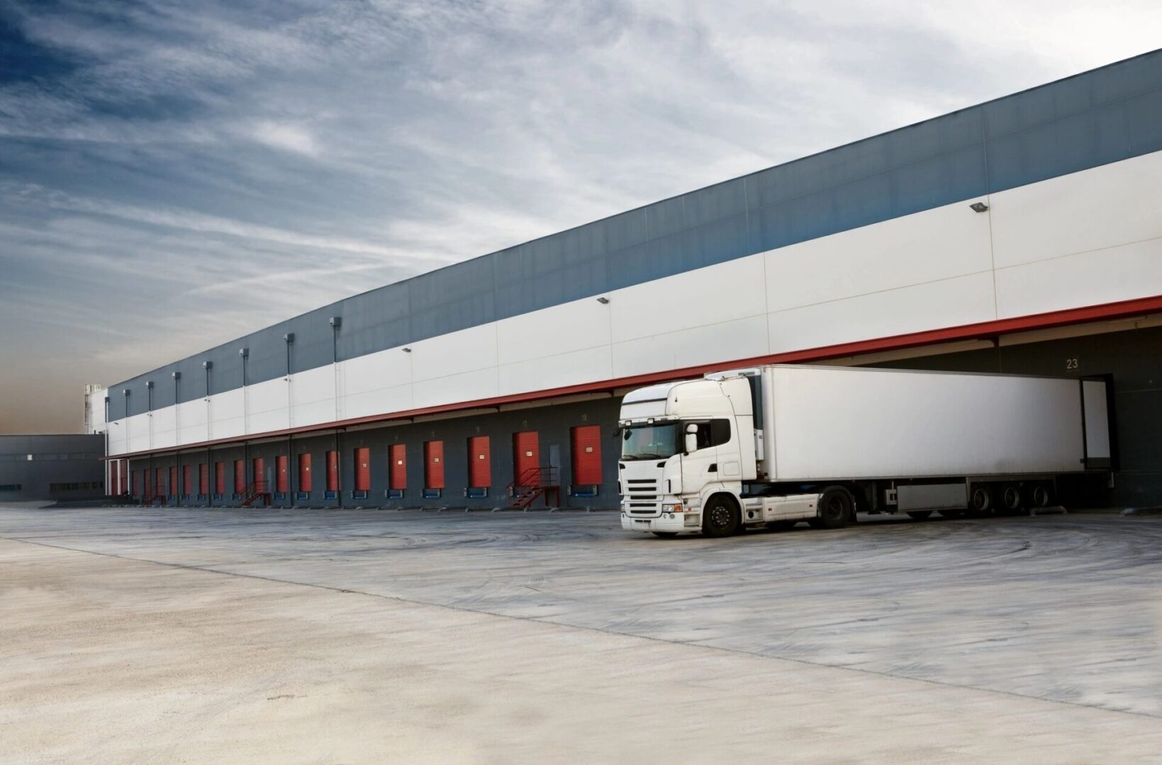 A large white truck parked at a warehouse loading dock under a cloudy sky.