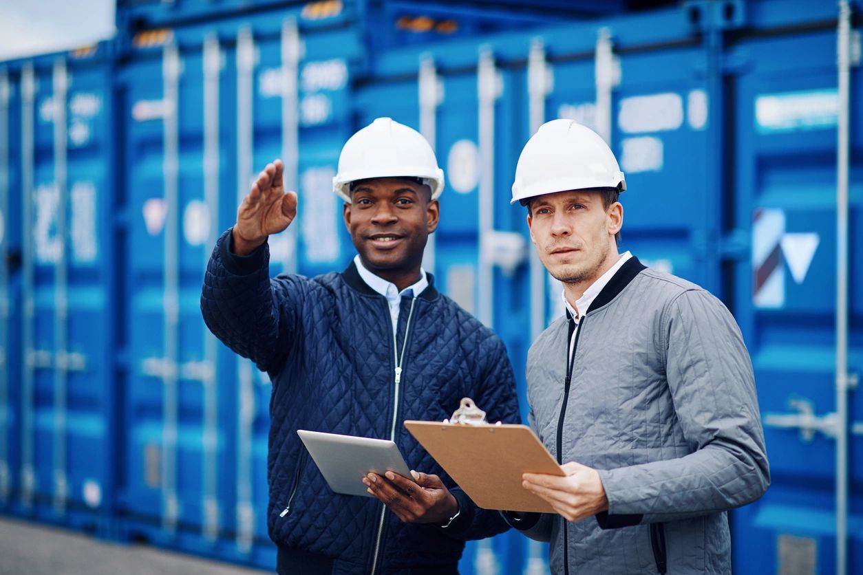 Two engineers in hard hats reviewing documents at a shipping yard.